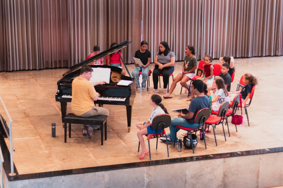 Aula coletiva de música em grupo, com professor ao piano e jovens sentados ao redor